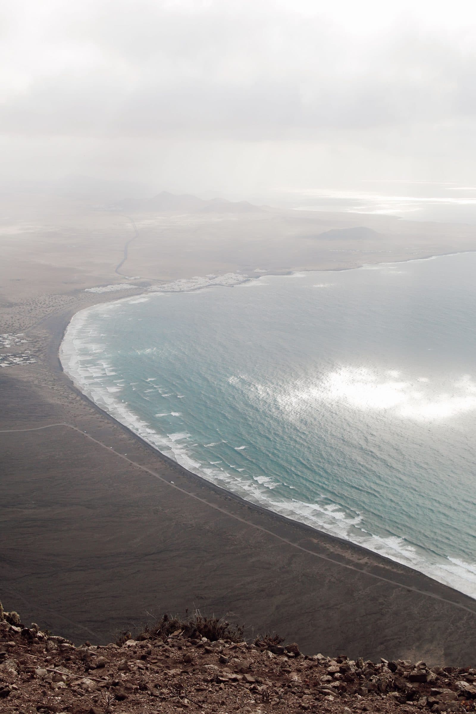 The long curve of Famara beach seen from above, where dark sand meets the surf.