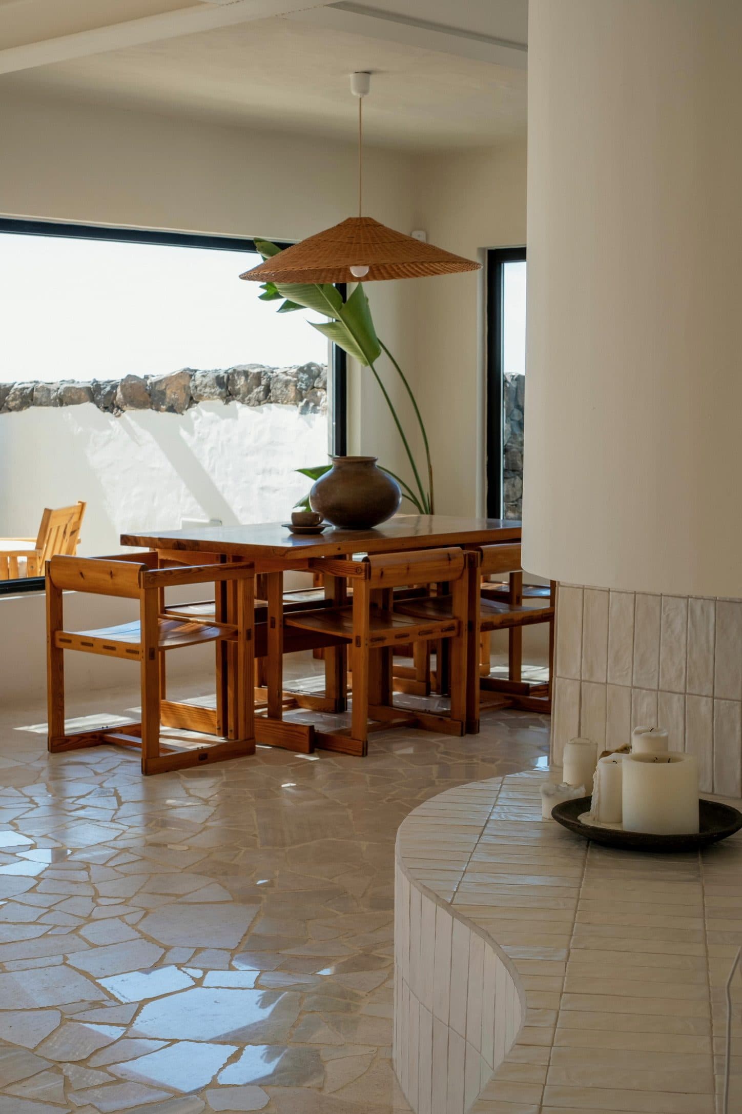 A timber dining table on a floor of irregular pale stone, beside a column of white plaster.