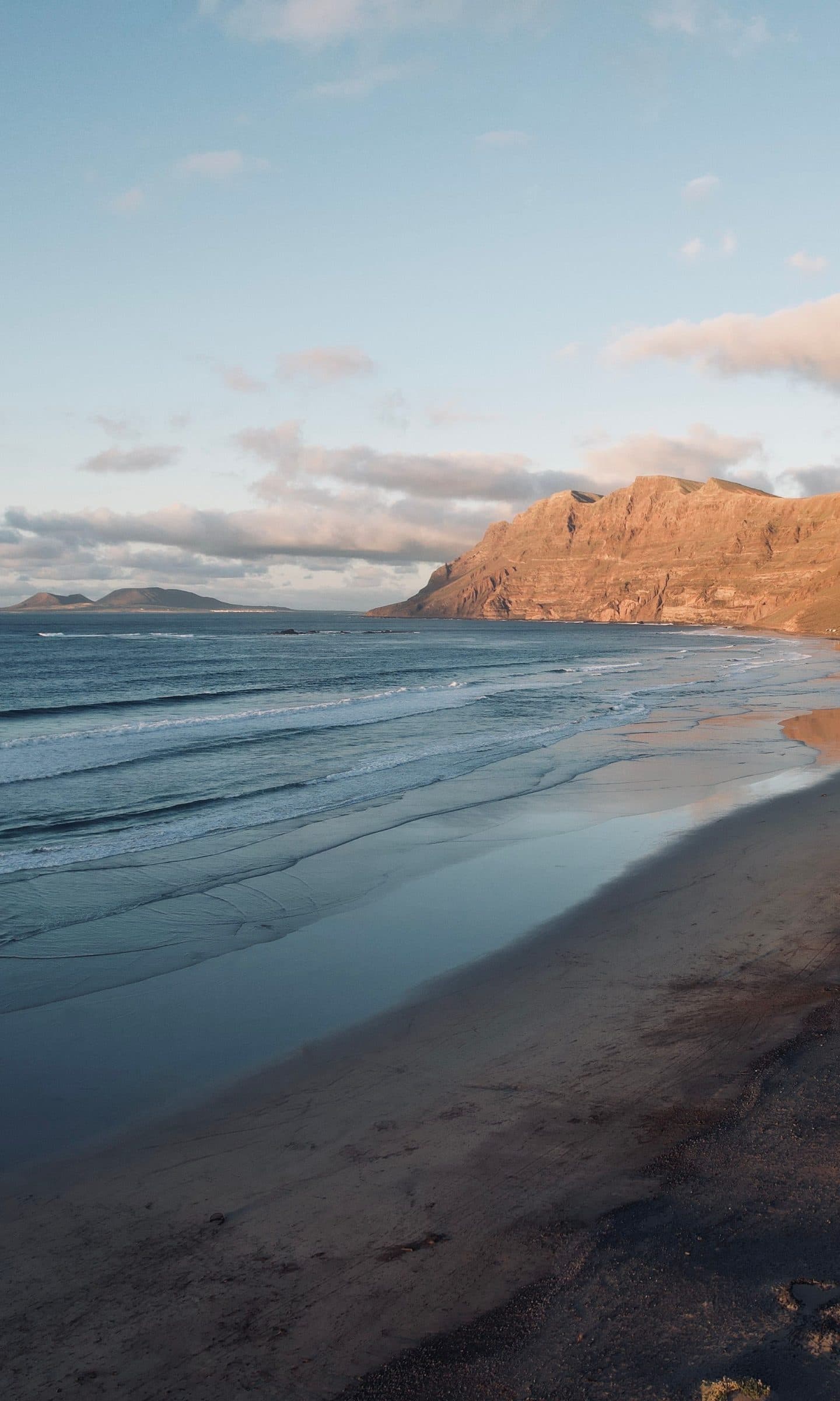 The Famara cliff glowing amber at sunset above a dark, wet beach.