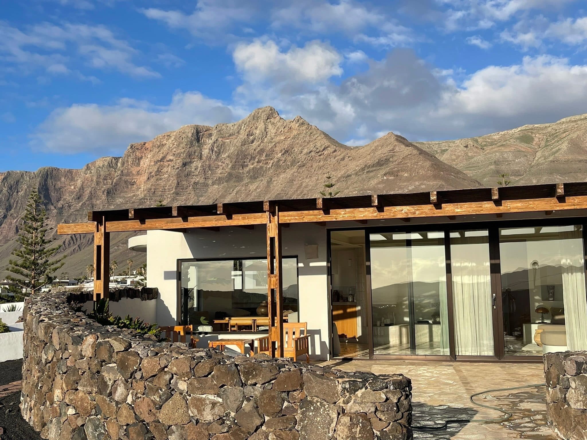 The suite in late golden light: a timber pergola and a curved basalt wall beneath the Famara cliffs.