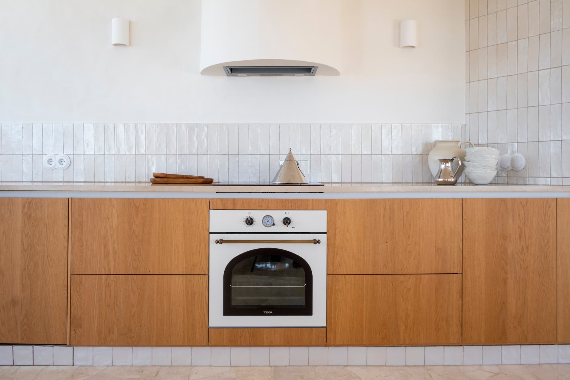 The kitchen in late golden light: timber cabinetry, white tile and a stone floor.